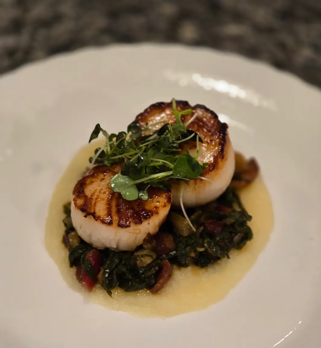 Golden seared scallops stacked over leafy greens with a pale sauce and fresh herbs.