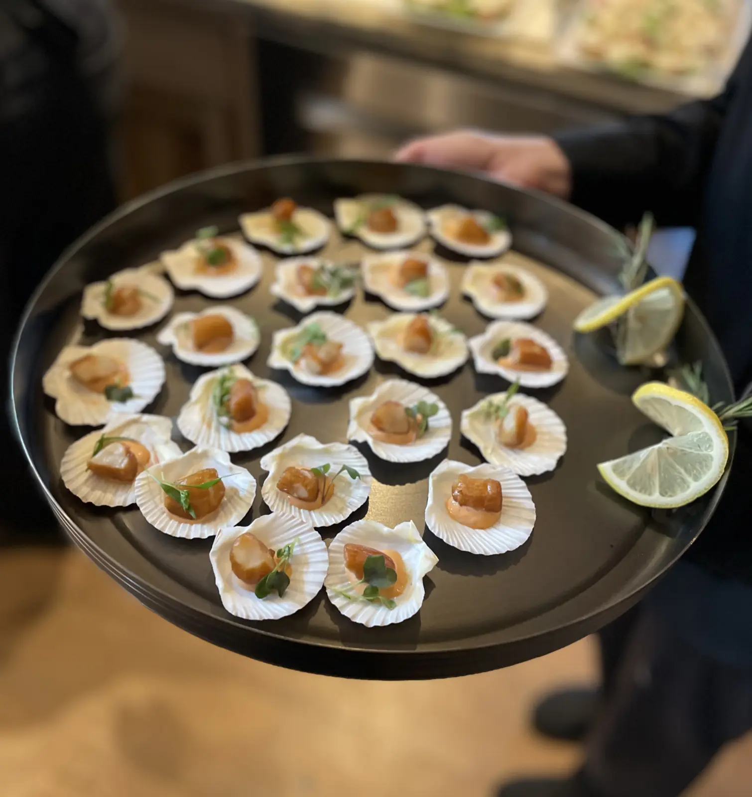 Smoked bay scallop appetizers served in shells on a round tray with lemon wedges.