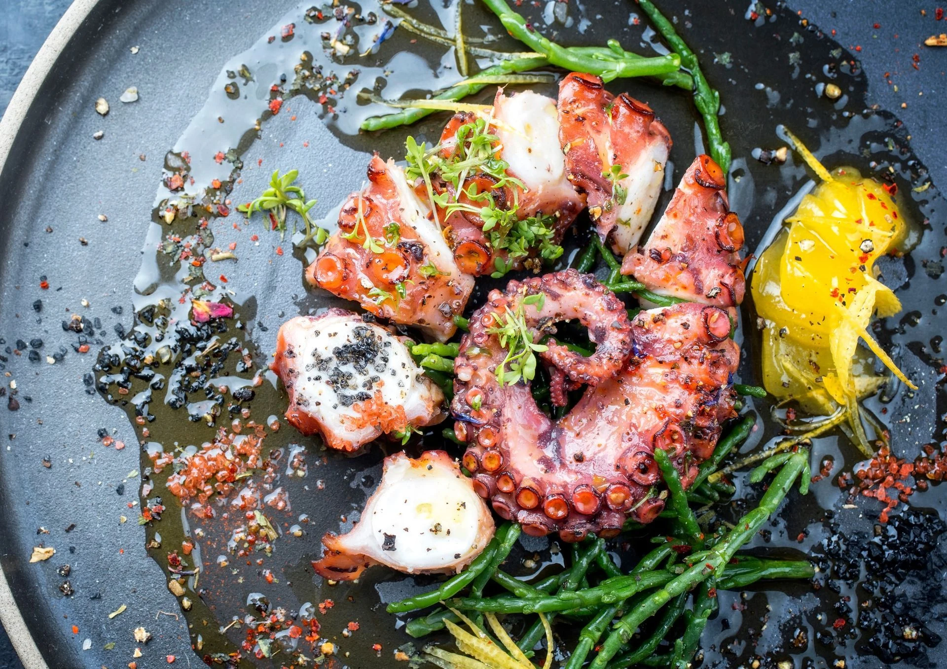 Grilled octopus with lemon and greens on a dark plate.